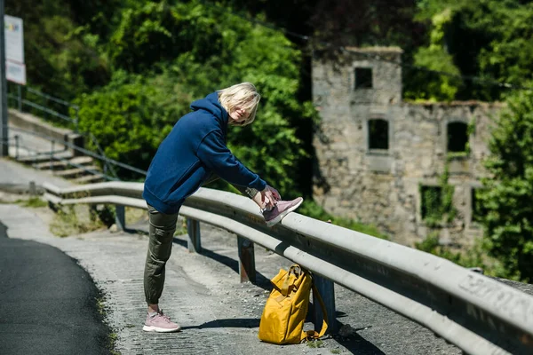 黄色のバックパックを持つ女性ハイカーは 道路上で彼女の靴ひもを結んでいます — ストック写真