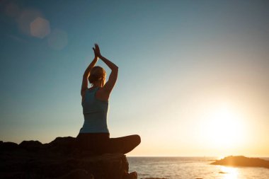 A woman does yoga, meditating in the lotus position on the ocean during sunset.   