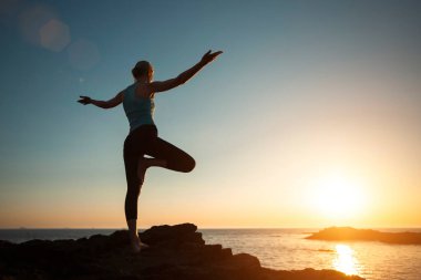 A woman does yoga, doing asana on the oceanfront during sunset.   