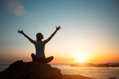 A woman does yoga in preparation for meditation near the ocean during sunset.   