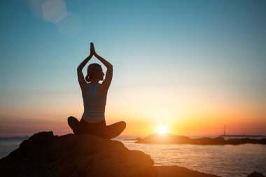 A woman of athletic build does yoga, meditating near the ocean at sunset.   