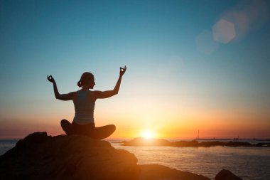 A middle-aged woman of athletic build does yoga, meditating on an ocean beach at dusk.   