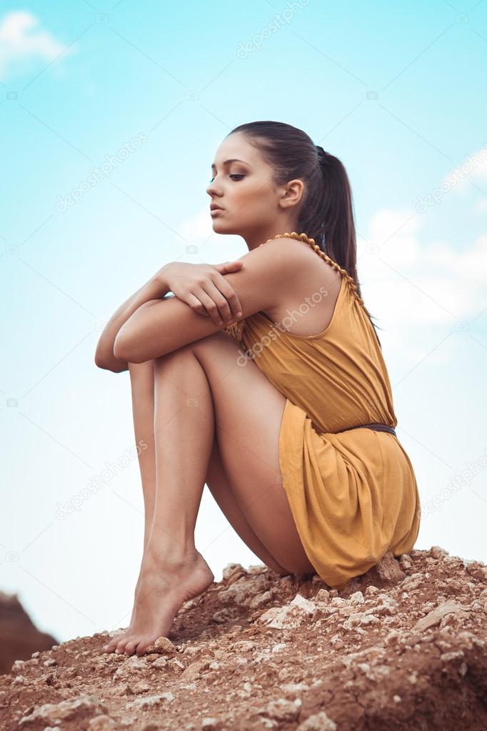 Beautiful brunette girl in yellow dress sitting on a rock