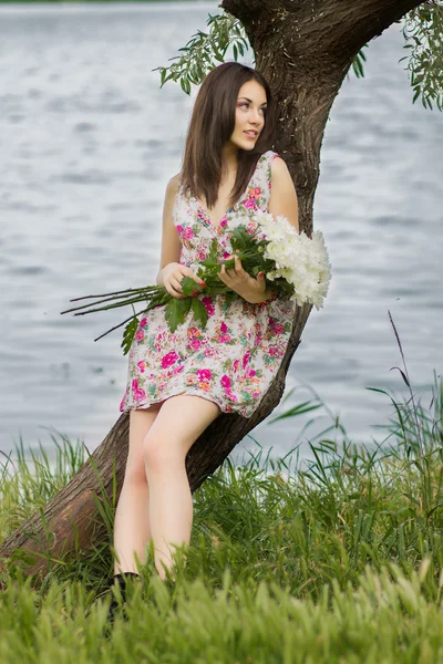 Hermosa joven morena de pie cerca de un árbol cerca de un río con un gran ramo de flores — Foto de Stock