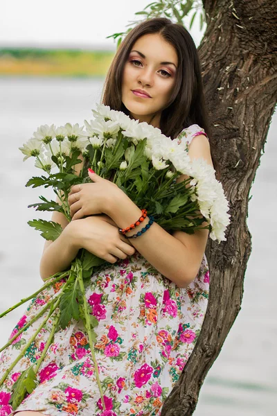 Όμορφη νεαρή γυναίκα μελαχρινή που στέκεται κοντά σε ένα δέντρο κοντά σε ένα ποτάμι με ένα μεγάλο μπουκέτο λουλούδια — Φωτογραφία Αρχείου