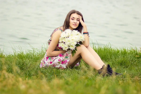 Beautiful young brunette woman sitting on the grass with a bouquet of flowers — Stock Photo, Image