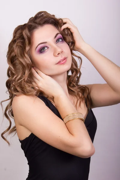 Retrato de una hermosa joven con el pelo rizado marrón en un vestido negro apretado y un brazalete de oro en su muñeca —  Fotos de Stock