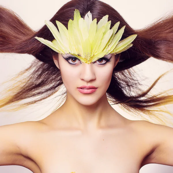 Portrait of beautiful young brown-haired woman with long hair in a studio. Makeup with long black arrow and yellow long feathers — Stock Photo, Image