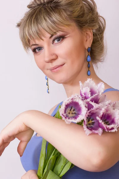 Egy szép fiatal szőke lány fehér háttéren egy kék ruhás portréja. nő pózol egy-egy csokor lila tulipán. — Stock Fotó