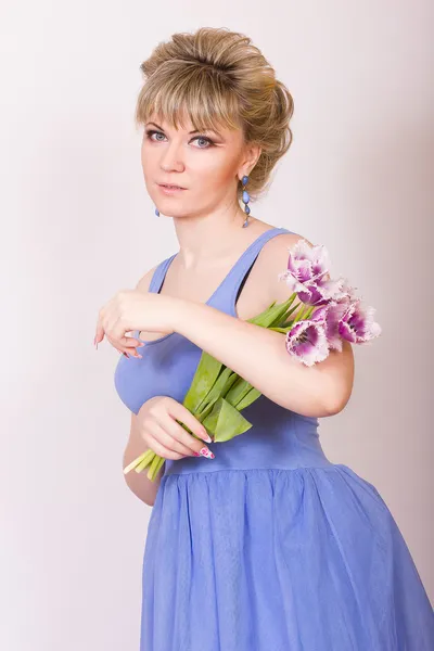 Retrato de uma bela jovem loira em um fundo branco em um vestido azul. Mulher posando com um buquê de tulipas roxas . — Fotografia de Stock