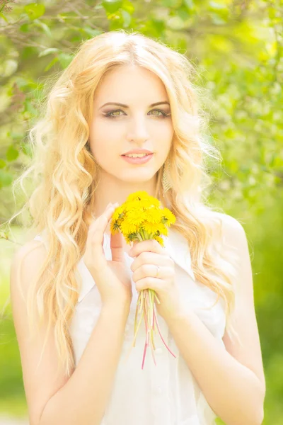 Retrato de una hermosa joven rubia con dientes de león. Chica posando en la naturaleza y sonriendo — Foto de Stock