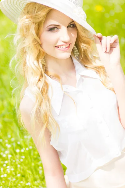 Retrato de una hermosa joven rubia con el pelo ondulado en la naturaleza. Chica en sombrero blanco sentado en la hierba en el parque —  Fotos de Stock