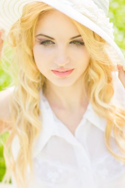 doğal dalgalı saçlı güzel bir genç sarışın kadın portresi. çim parkta oturan beyaz şapkalı kız