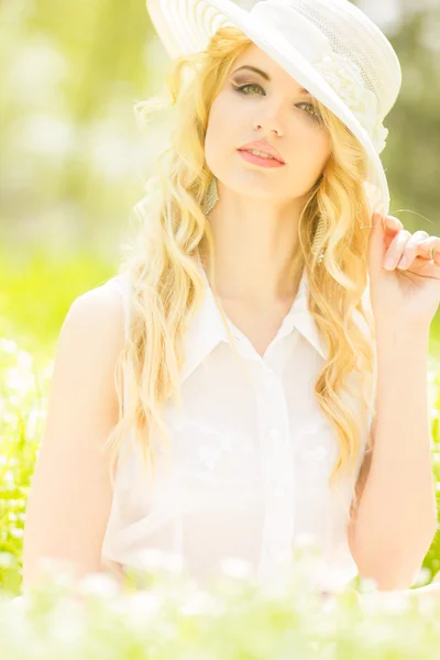 Retrato de uma bela jovem loira com cabelo ondulado na natureza. Menina de chapéu branco sentado na grama no parque — Fotografia de Stock