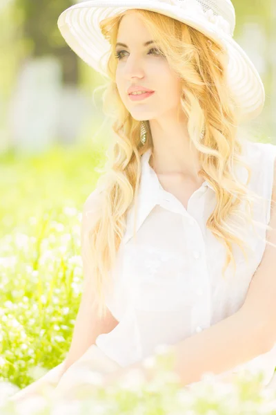 Portret van een mooie jonge blonde vrouw met golvend haar in de natuur. meisje in witte hoed zittend op het gras in het park — Stockfoto