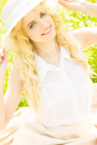 Retrato de uma bela jovem loira com cabelo ondulado na natureza. Menina de chapéu branco sentado na grama no parque — Fotografia de Stock