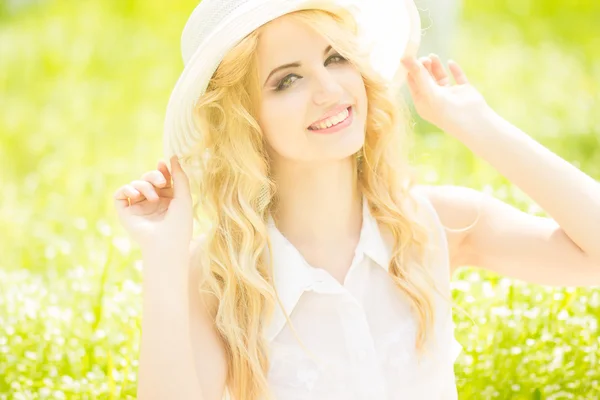 自然の中でウェーブのかかった髪と美しい若いブロンドの女性の肖像画。公園の芝生の上に座っている白い帽子の少女 — ストック写真