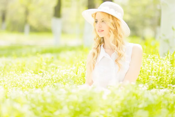 Portrait of a beautiful young blonde woman with wavy hair in nature. Girl in white hat sitting on the grass in the park — Stock Photo, Image