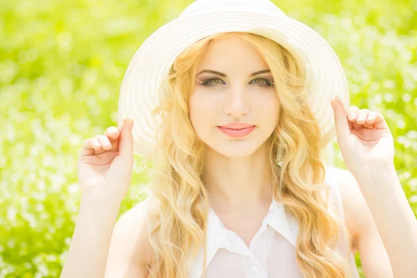 Portret van een mooie jonge blonde vrouw met golvend haar in de natuur. meisje in witte hoed zittend op het gras in het park — Stockfoto