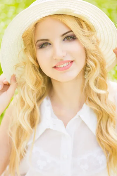 Retrato de una hermosa joven rubia con el pelo ondulado en la naturaleza. Chica en sombrero blanco sentado en la hierba en el parque —  Fotos de Stock