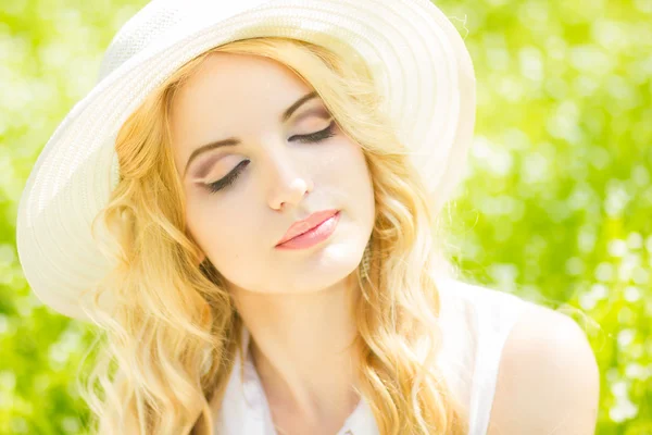 Ritratto di una bella giovane donna bionda dai capelli ondulati in natura. Ragazza in cappello bianco seduta sull'erba nel parco — Foto Stock