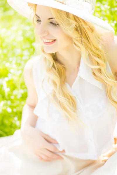 Retrato de una hermosa joven rubia con el pelo ondulado en la naturaleza. Chica en sombrero blanco sentado en la hierba en el parque — Foto de Stock