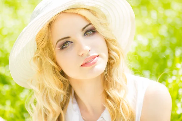Retrato de una hermosa joven rubia con el pelo ondulado en la naturaleza. Chica en sombrero blanco sentado en la hierba en el parque —  Fotos de Stock