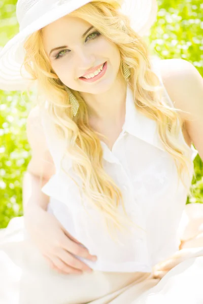 Retrato de una hermosa joven rubia con el pelo ondulado en la naturaleza. Chica en sombrero blanco sentado en la hierba en el parque — Foto de Stock