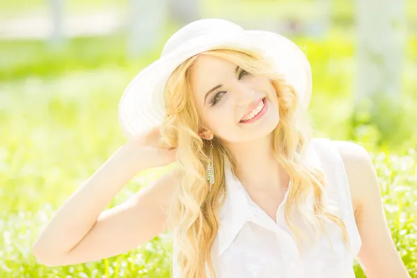 Retrato de una hermosa joven rubia con el pelo ondulado en la naturaleza. Chica en sombrero blanco sentado en la hierba en el parque —  Fotos de Stock