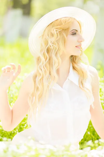Retrato de una hermosa joven rubia con el pelo ondulado en la naturaleza. Chica en sombrero blanco sentado en la hierba en el parque —  Fotos de Stock