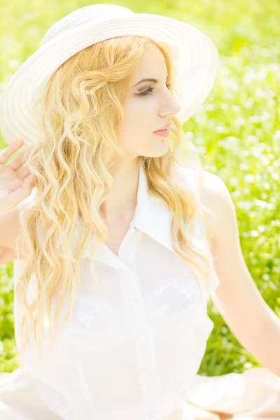 自然の中でウェーブのかかった髪と美しい若いブロンドの女性の肖像画。公園の芝生の上に座っている白い帽子の少女 — ストック写真
