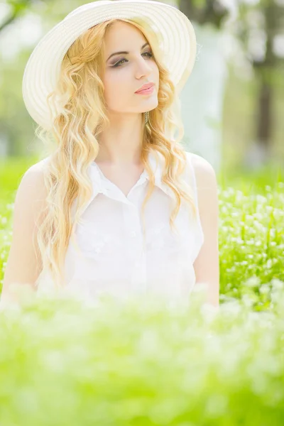 Retrato de una hermosa joven rubia con el pelo ondulado en la naturaleza. Chica en sombrero blanco sentado en la hierba en el parque — Foto de Stock