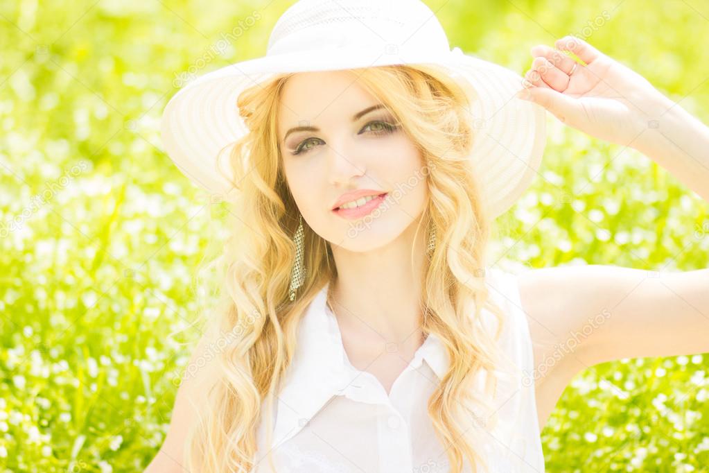 Portrait of a beautiful young blonde woman with wavy hair in nature. Girl in white hat sitting on the grass in the park