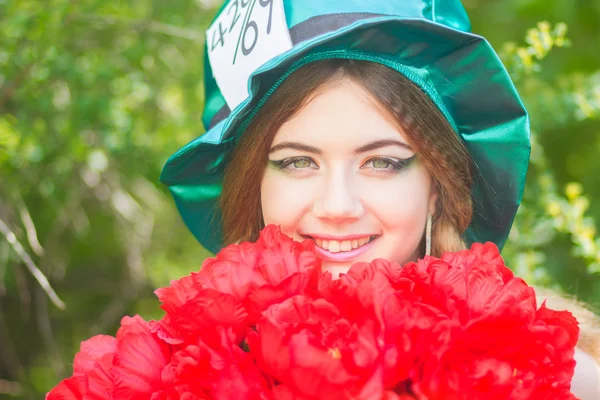 Porträt einer schönen jungen Frau im Kostüm des verrückten Hutmachers in der Natur. Mädchen posiert mit einem Strauß roter Pfingstrosen — Stockfoto