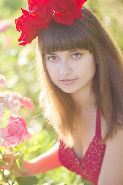 Porträtt av en vacker ung kvinna med brunt hår i rött ljus klänning. flicka i trädgården med rosenbuskar — Stockfoto