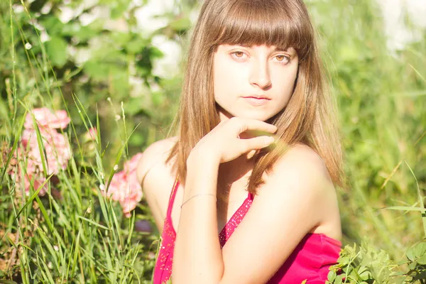 茶色の髪赤い光のドレスで美しい若い女性の肖像画。バラの茂みと庭の女の子 — ストック写真