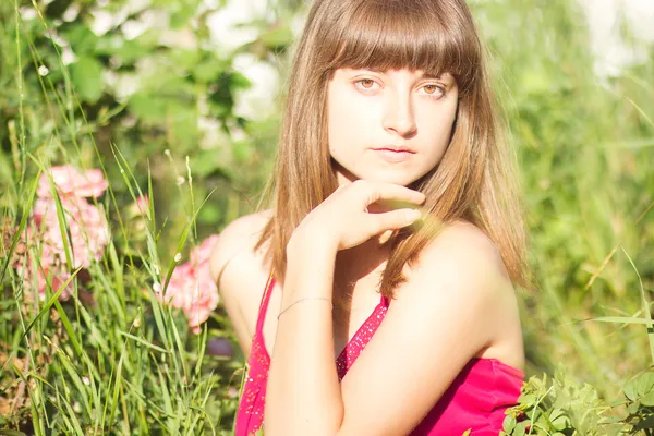 Ritratto di una bella giovane donna dai capelli castani in abito rosso chiaro. Ragazza in giardino con cespugli di rose — Foto Stock