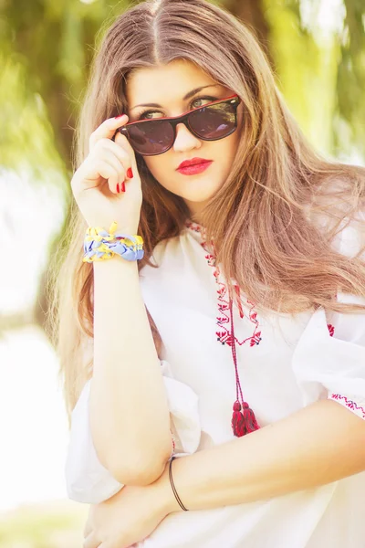 Portrait of a beautiful young woman with long brown hair who poses with sunglasses. The girl is on the nature and smiling. — Stock Photo, Image