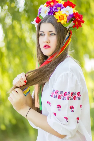 Portret to piękna młoda kobieta z długie brązowe włosy w przyrodzie w bluzka. dziewczyna pozuje z wieniec kolorowych kwiatów. nieostrość. — Zdjęcie stockowe