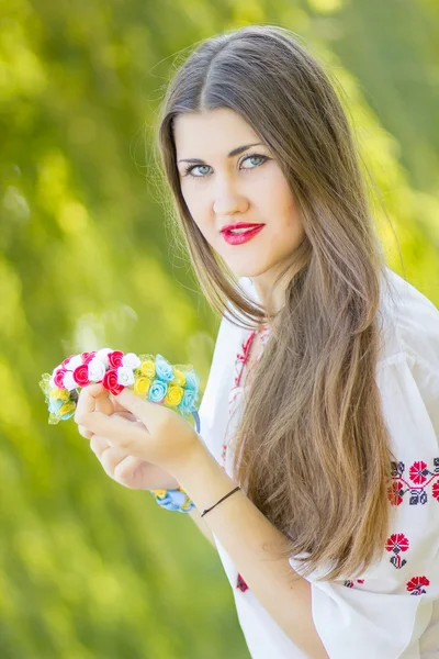 Portrait of a beautiful young woman with long brown hair in nature in a blouse. Girl posing with a wreath of colorful flowers. Soft focus. — Stock Photo, Image