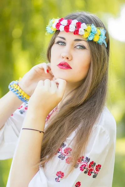 Retrato de uma bela jovem com longos cabelos castanhos na natureza em uma blusa. Menina posando com uma coroa de flores coloridas. Foco suave . — Fotografia de Stock