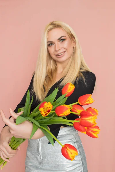 Fashion model woman in fashionable clothes on pink background. Wearing stylish clothing, black blouse, silver skirt. Posing in studio. Holding red tulips in her hands
