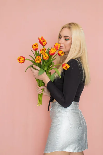 Fashion model woman in fashionable clothes on pink background. Wearing stylish clothing, black blouse, silver skirt. Posing in studio. Holding red tulips in her hands