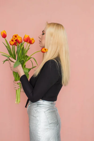 Fashion model woman in fashionable clothes on pink background. Wearing stylish clothing, black blouse, silver skirt. Posing in studio. Holding red tulips in her hands