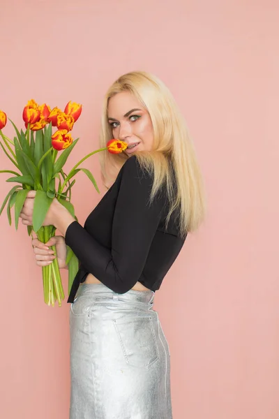 Fashion model woman in fashionable clothes on pink background. Wearing stylish clothing, black blouse, silver skirt. Posing in studio. Holding red tulips in her hands