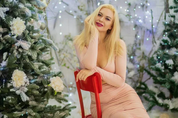 迷人的金发女人穿着米色性感的衣服坐在红色的椅子上 背对着圣诞树 圣诞及新年照片 — 图库照片