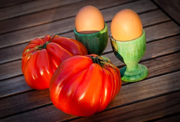 Två blanka mogen biff tomat och två ägg i grönt trä äggkoppar isolerad på trä bakgrund. Gyllene ljus och lång kväll skuggor. — Stockfoto