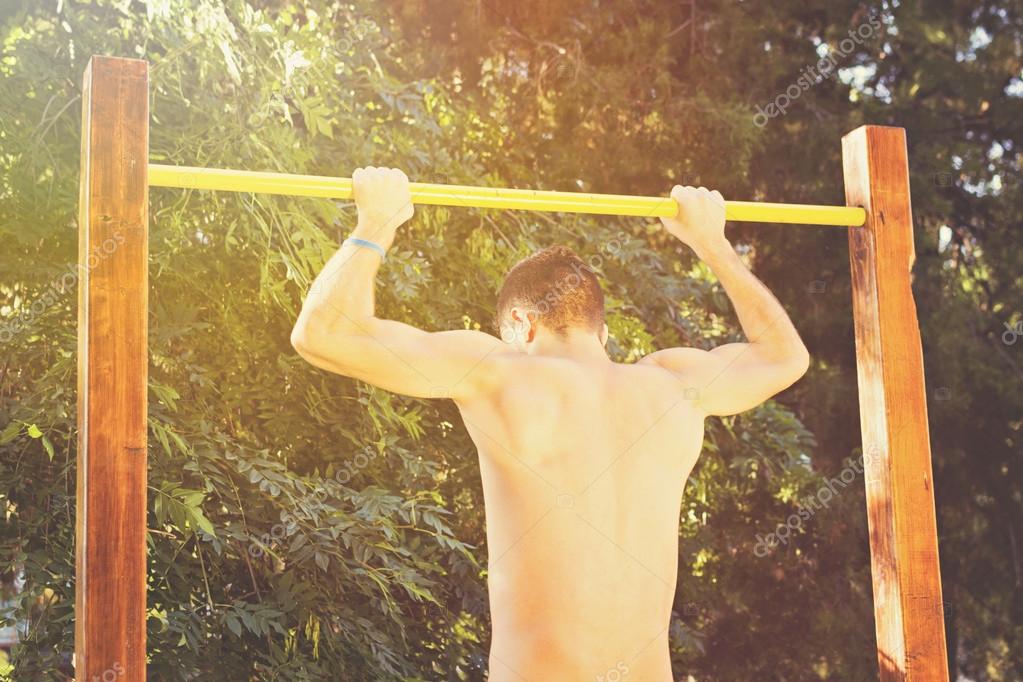 Closeup shot of a young Caucasian muscular topless man doing pull-ups on ba...