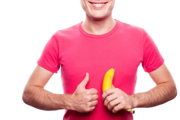 Smiling handsome guy holding yellow banana and showing ok (thumb up) near his stomach over white background. studio shot. — Stock Photo, Image