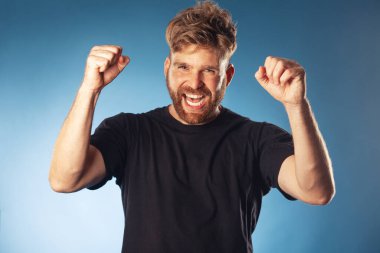 Spor zaferi konsepti. Çığlık atan karizmatik, aktif, 30 yaşında, mavi arka planda elleri havada poz veren birinin duygusal portresi. Mükemmel saç kesimi. Hippi tarzı. Beyaz, parlak bir gülümseme. Stüdyo çekimi